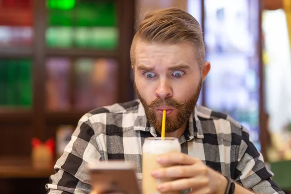 Σοκαρισμένος νεαρός άντρας που πίνει κακάο από άχυρο και κοιτάζει μηνύματα στα κοινωνικά δίκτυα ενώ κάθεται σε ένα τραπέζι σε ένα καφέ. Η έννοια της συνομιλίας και των κοινωνικών δικτύων. — Φωτογραφία Αρχείου