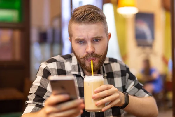 Pensativo joven y elegante joven hipster hombre con bigote y barba está buscando en la red social en el teléfono inteligente utilizando Internet de alta velocidad gratuita. Concepto de comunicación en las redes sociales . — Foto de Stock