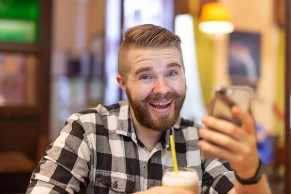 Un joven elegante con bigote y barba está viendo una red social usando un teléfono inteligente mientras está sentado en un café un fin de semana. El concepto de dependencia de las redes sociales . — Foto de Stock