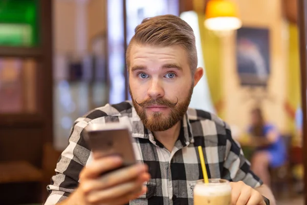 Retrato de un joven apuesto y reflexivo con bigote y barba mirando a la cámara en un café sobre un fondo borroso. El concepto de estudiante o los hombres con estilo . — Foto de Stock