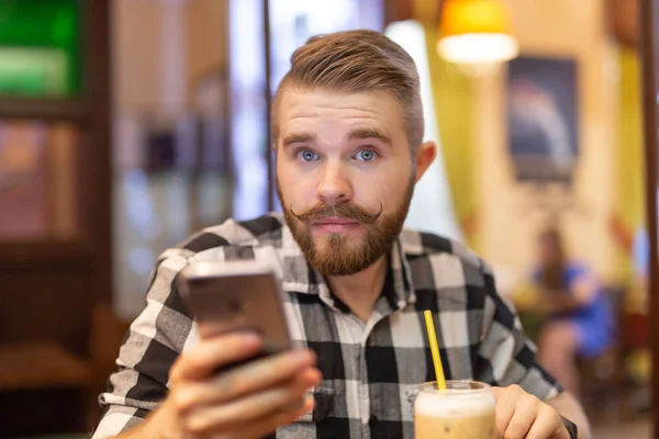 Pensativo joven y elegante joven hipster hombre con bigote y barba está buscando en la red social en el teléfono inteligente utilizando Internet de alta velocidad gratuita. Concepto de comunicación en las redes sociales . — Foto de Stock