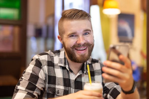 Un joven elegante con bigote y barba está viendo una red social usando un teléfono inteligente mientras está sentado en un café un fin de semana. El concepto de dependencia de las redes sociales . — Foto de Stock