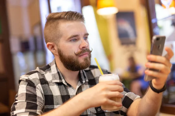 Un joven elegante con bigote y barba está viendo una red social usando un teléfono inteligente mientras está sentado en un café un fin de semana. El concepto de dependencia de las redes sociales . — Foto de Stock