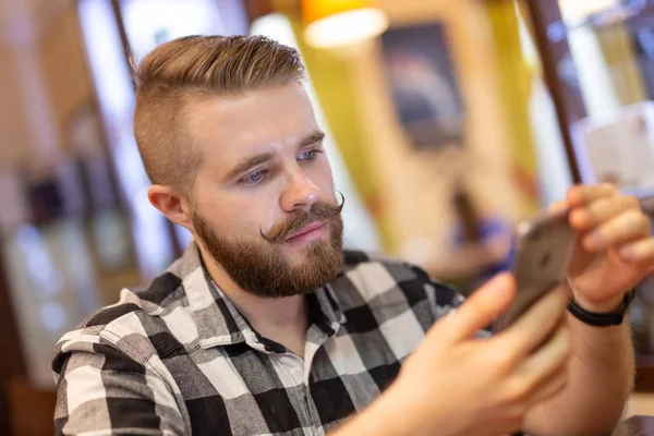 Un joven elegante con bigote y barba está viendo una red social usando un teléfono inteligente mientras está sentado en un café un fin de semana. El concepto de dependencia de las redes sociales . — Foto de Stock