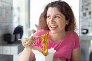 Bir kafede Çin eriştesi yiyen ve pencereden dışarı bakan genç ve neşeli bir kızın portresi. Sağlıklı Asya mutfağı kavramı.