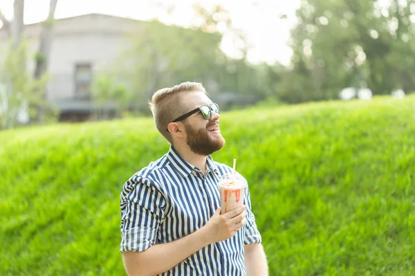 Zijaanzicht van een stijlvolle jongeman met een baard die een milkshake vasthoudt en het uitzicht op de stad bewondert terwijl je op een warme zomerdag in het Park wandelt. Het concept van rust en ontspanning. — Stockfoto