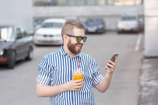 Vista lateral de un joven y elegante hombre hipster con gafas y un bigote y jugo en sus manos charlando en las redes sociales usando un teléfono inteligente e internet inalámbrico en un cálido día de verano . — Foto de Stock