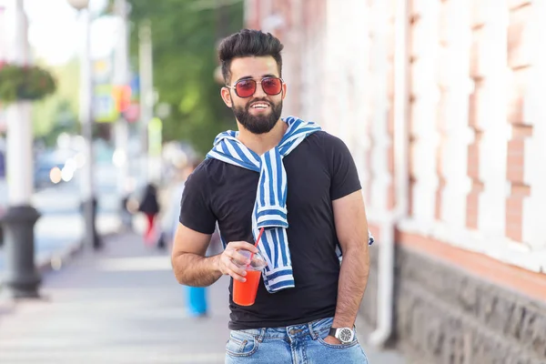 Guapo joven estudiante árabe alegre con un bigote elegante y una barba con jugo en las manos, caminando por la ciudad después de un día de trabajo. Concepto de positivo y descanso . — Foto de Stock