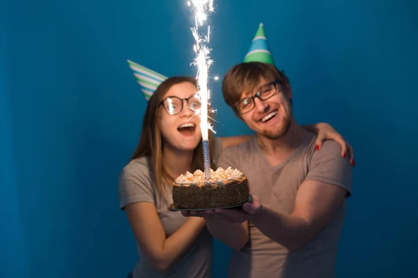 Funny Nerd mężczyzna i kobieta noszą wakacje czapki i okulary gospodarstwa tort urodzinowy z brylant na niebieskim tle — Zdjęcie stockowe