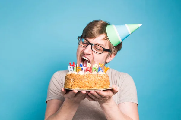 Divertente maschio in carta cappello di congratulazione cercando di mordere una torta con un felice compleanno candele stand su uno sfondo blu . — Foto Stock