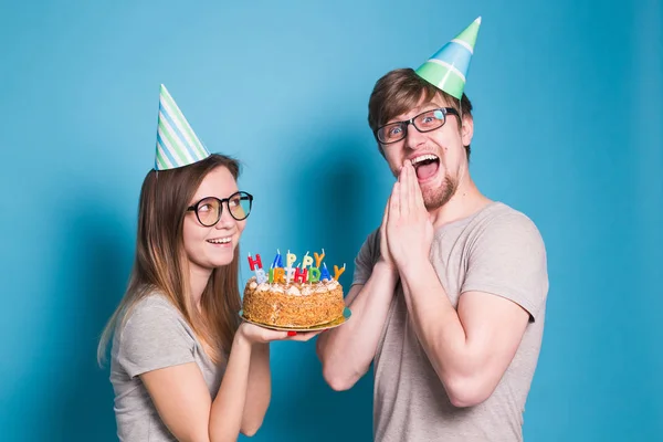 Divertido nerd hombre y mujer están usando días festivos gorras y gafas celebración de pastel de cumpleaños con velas sobre fondo azul — Foto de Stock