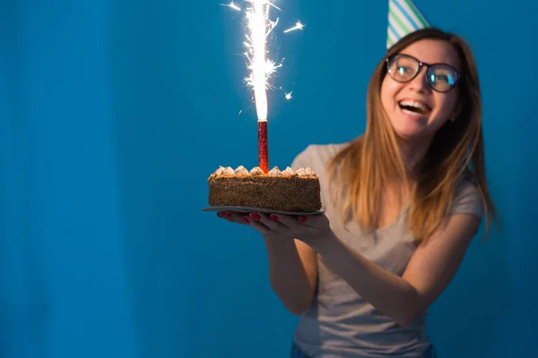 Wesoły młody niewyraźne uczennice w okularach trzyma tort gratulacyjny ze świecą stojącą na niebieskim tle. Koncepcja urodzin. — Zdjęcie stockowe