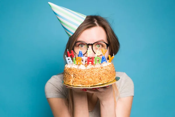 Close up funny positive girl in glasses and greeting paper hat holding a happy birthday cake in her hands standing on a blue background. Royalty Free Stock Images