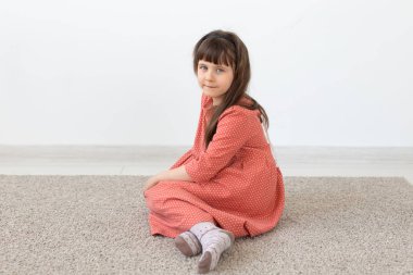 Küçük sevimli kız beyaz bir duvara karşı mütevazı uzun pembe elbise ile oturuyor. Küçük masum çocuklar kavramı.