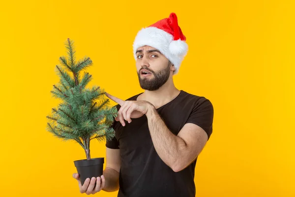 Joven guapo sosteniendo una caja de regalo roja y un árbol de Navidad en sus manos posando en una gorra de Año Nuevo sobre un fondo amarillo. Feliz Navidad y Feliz Año Nuevo concepto de felicitaciones . — Foto de Stock