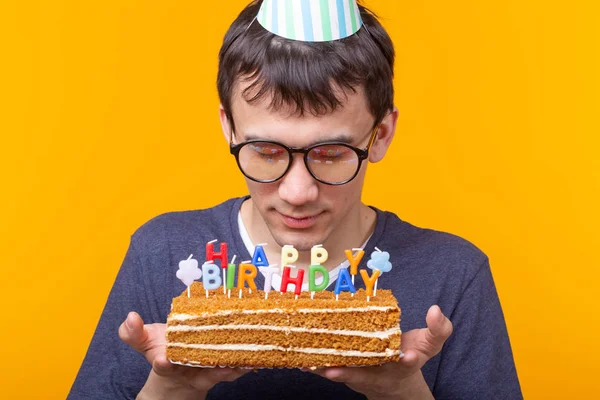 Close-up de cara positivo engraçado em óculos segura em suas mãos um bolo caseiro com a inscrição feliz aniversário posando em um fundo amarelo. Conceito de feriados e aniversários . — Fotografia de Stock