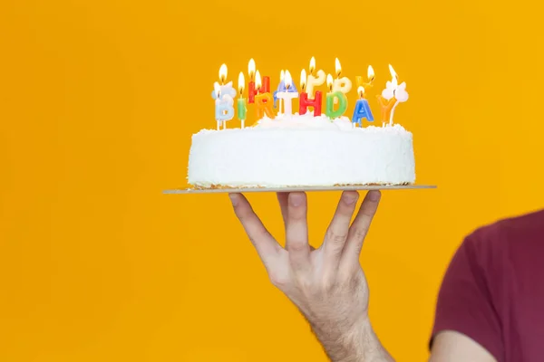 Mãos que mantêm um bolo de aniversário com velas e o aniversário de inscrição em um contexto amarelo. Parabéns pelo aniversário e aniversário. Espaço de cópia . — Fotografia de Stock