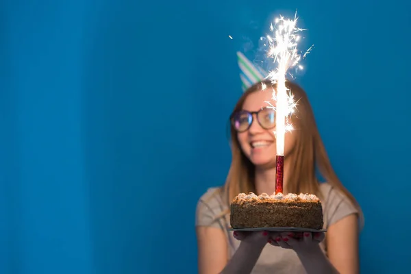 Χαρούμενος νέος θολή κορίτσι μαθητής σε γυαλιά κρατώντας ένα συγχαρητήριο κέικ με ένα κερί που στέκεται σε μπλε φόντο. Ιδέα γενεθλίων. — Φωτογραφία Αρχείου