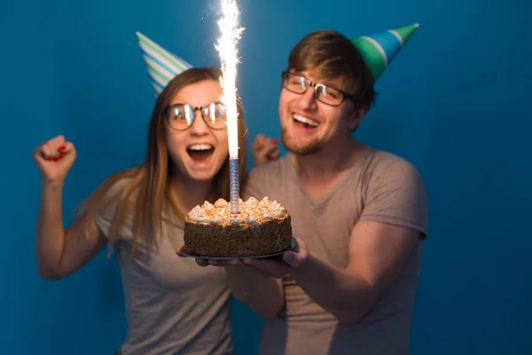 Funny young couple in paper caps and with a cake make a foolish face and wish happy birthday while standing against a blue background. Concept of congratulations and fooling around. — Stock Photo, Image