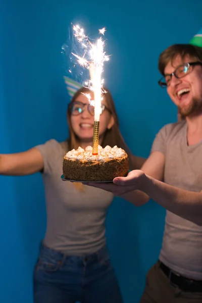 Веселая молодая пара очаровательный парень и милая девушка в бумажных шляпах делают глупое лицо и держать в руках торт день рождения стоя на синем фоне . — стоковое фото