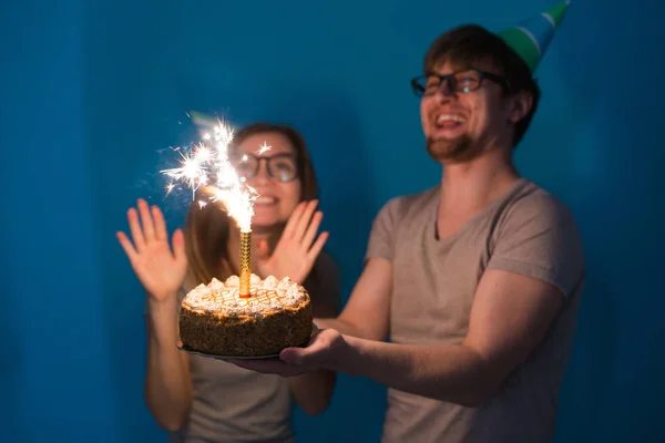 Jóvenes estudiantes alegres encantadora chica y buen chico en gorras de papel de saludo sosteniendo un pastel con una vela chispas de bengala. Concepto de felicitación por el cumpleaños y aniversario —  Fotos de Stock