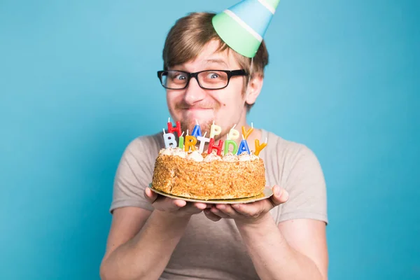 Estudante jovem louco engraçado em um chapéu de saudação se alegra ao lado do bolo com a inscrição feliz aniversário de pé em um fundo azul. Conceito de saudações. Espaço de cópia — Fotografia de Stock