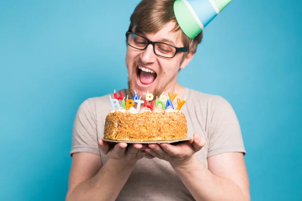 Lustiger Mann mit Gratulationsmütze aus Papier, der versucht, eine Torte mit einem Happy Birthday Kerzen abzubeißen stehen auf blauem Hintergrund. — Stockfoto