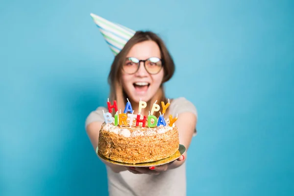 Menina engraçada louca em um chapéu de papel e óculos segurando um grande bolo de aniversário no fundo azul. Conceito de brincadeira e saudações . — Fotografia de Stock