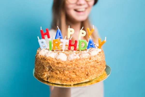 Szalona zabawna dziewczyna w papierowym kapeluszu i okularach posiadających duży tort urodzinowy na niebieskim tle. Koncepcja Prank i pozdrowienia. — Zdjęcie stockowe