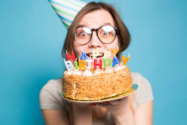 Közelről vicces pozitív lány a szemüveg és üdvözlő papír kalap gazdaság boldog születésnapi torta a kezében állt a kék háttér. — Stock Fotó