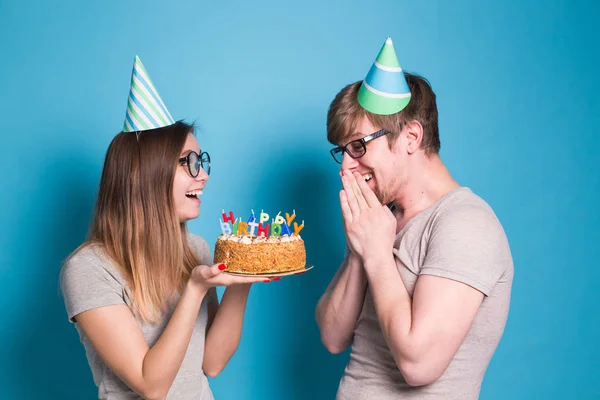 Neşeli genç çift sevimli adam ve kağıt şapkalı sevimli kız aptal yüz yapmak ve ellerinde mavi bir arka plan üzerinde duran bir doğum günü pastası tutun. — Stok fotoğraf