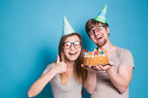 Funny Nerd mężczyzna i kobieta noszą wakacje czapki i okulary gospodarstwa tort urodzinowy ze świecami na niebieskim tle — Zdjęcie stockowe