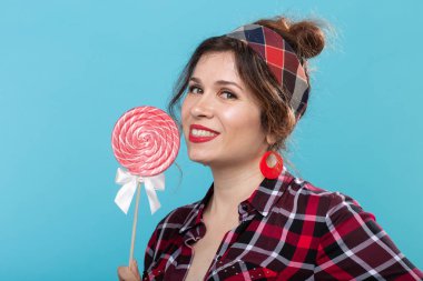 Elinde renkli lolipop tutan ve mavi arka planda poz birini yalayan retro giysiler içinde Büyüleyici genç kadın. Tatlılar ve tatiller için aşk kavramı.