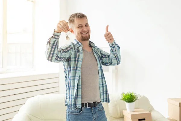 Jovem bonitão mostra as chaves de seu novo apartamento e mostrando os polegares para cima enquanto está na sala de estar. Conceito de hipoteca e empréstimo de casa para uma família jovem — Fotografia de Stock