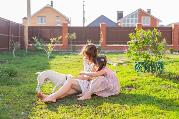 Мила молода мати і дочка сидять на траві свого заміського будинку босоніж поруч зі своїм улюбленим собакою в сонячний літній день. Концепція сімейного відпочинку . — стокове фото