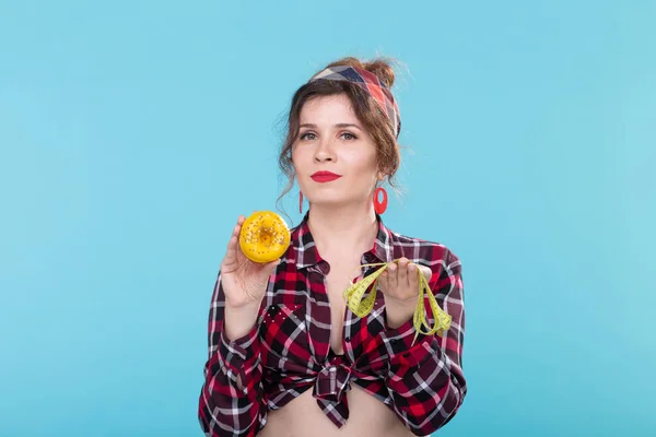 Close-up surprised young beautiful woman looking at measuring tape and holding a green donut in hands posing on a blue background. Concept of diet and rejection of harmful high-calorie foods.
