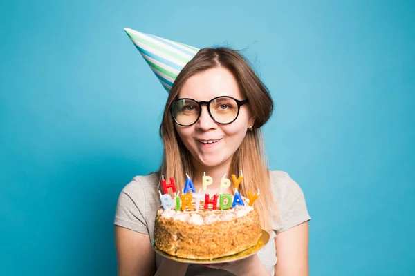 파란색 배경에 큰 생일 케이크를 들고 종이 모자와 안경에 미친 재미 소녀. 장난과 인사의 개념. — 스톡 사진