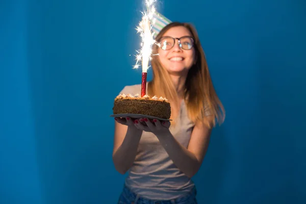 Joven estudiante alegre borrosa en gafas sosteniendo un pastel de felicitación con una vela de pie sobre un fondo azul. Concepto de cumpleaños . — Foto de Stock