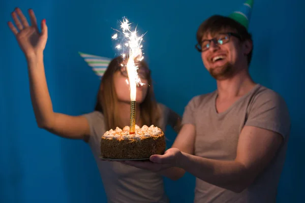 欢快的年轻夫妇迷人的家伙和可爱的女孩在纸帽子使愚蠢的脸,并举行在他们的手中的生日蛋糕站在蓝色背景. — 图库照片