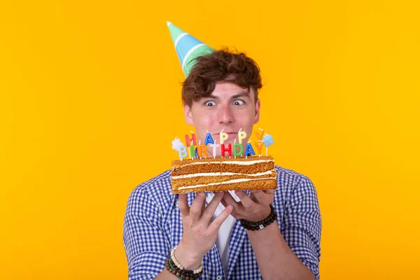 Kağıt kutlama şapka deli neşeli genç adam sarı bir arka plan üzerinde duran kek mutlu doğum günü tutan. Jubilee tebrikler kavramı. — Stok fotoğraf