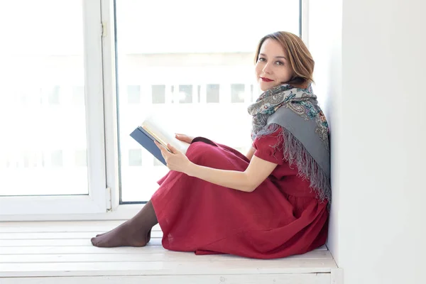 Boční pohled na okouzlující mladou dívku v dlouhém skromném oděvu a šátek čte knihu, zatímco sedí na okenní římse. Koncept mládí a romantiky. — Stock fotografie