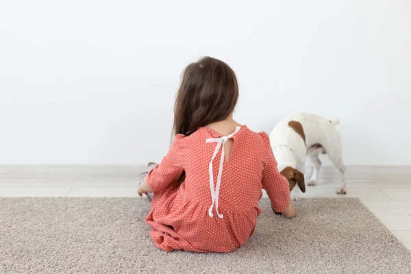 흰색 배경에 그녀의 사랑하는 개 잭 러셀 테리어 옆에 바닥에 앉아 빨간색 물방울 무늬 드레스에 어린 소녀의 뒷모습. 어린이 제품의 개념. — 스톡 사진