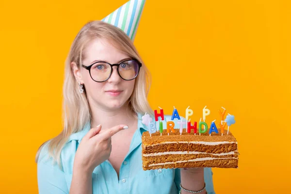 노란색 배경에 그녀의 손에 축하 케이크를 들고 종이 모자와 안경 재미있는 긍정적 인 소녀의 초상화. 개념과 재미와 축하. 광고 공간. — 스톡 사진