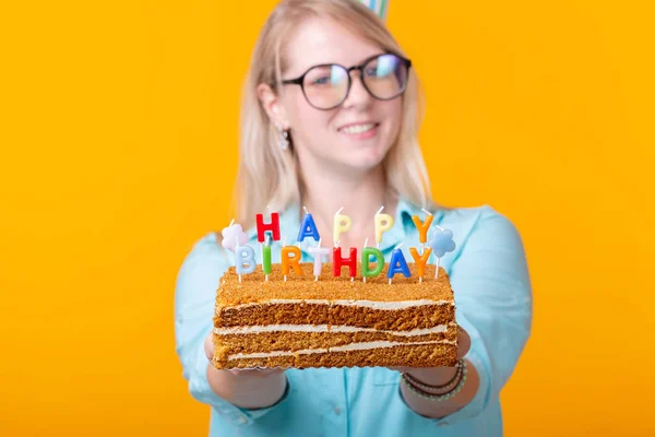 Jovem alegre louca no papel chapéu congratulatório segurando bolos feliz aniversário de pé em um fundo amarelo. Conceito de parabéns Jubileu . — Fotografia de Stock