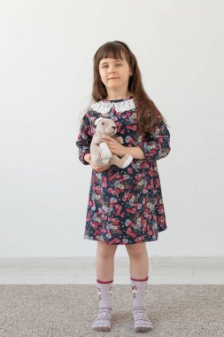 Çiçek elbiseli küçük sevimli kız, beyaz bir arka plan üzerinde duran en sevdiği oyuncak tavşanı nazikçe kucaklıyor. Çocuk oyuncakları ve ürünleri kavramı