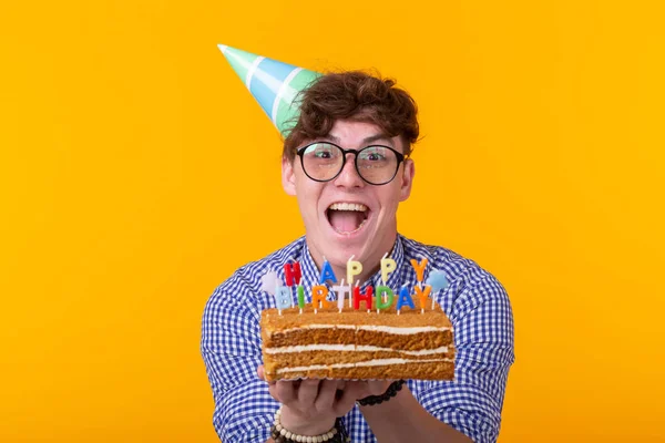 Loco joven alegre en papel sombrero de felicitación celebración tortas feliz cumpleaños de pie sobre un fondo amarillo. Concepto de felicitaciones jubilar . —  Fotos de Stock