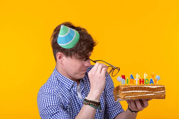 Jovem alegre louco no papel chapéu congratulatório segurando bolos feliz aniversário de pé em um fundo amarelo. Conceito de parabéns Jubileu . — Fotografia de Stock