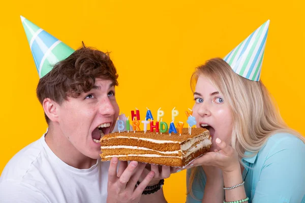 Close up de bizarro jovem casal menina e menino em chapéus de papel quer morder um pedaço de bolo de parabéns. Conceito de aniversário e parabéns . — Fotografia de Stock