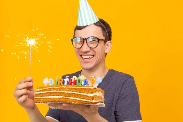 Portré egy vicces pozitív fickó egy papír sapkát és szemüveg tartó gratuláló házi torta a kezében egy sárga háttér. Koncepció és szórakozás és ünneplés. — Stock Fotó