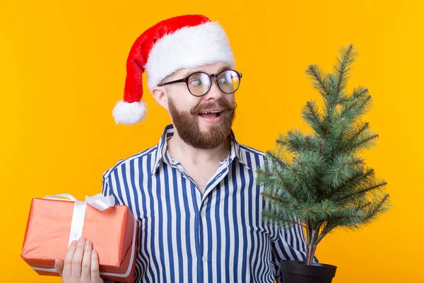 Positieve jonge man Kerstman met geschenken en kerstboom poseren op een gele achtergrond. Kerst en Nieuwjaar vakantie concept. — Stockfoto